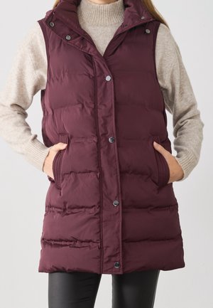 Vrouw draagt een mouwloos bordeaux puffer-vestje met drukknopen over een beige trui, handen in de zakken van het vest.