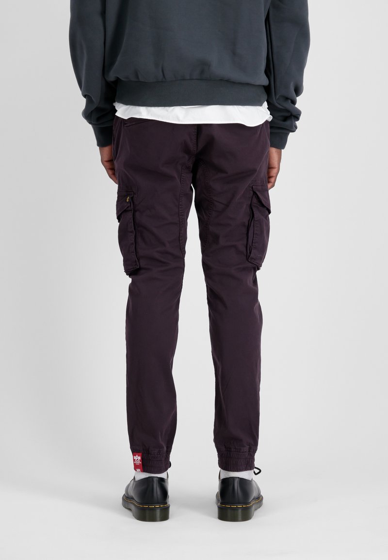 Fioletowe spodnie cargo z bocznymi kieszeniami, elastycznymi mankietami i czerwoną naszywką z logo na dole, wykonane z lekkiego materiału.