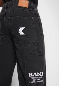 Svarta denimbyxor med broderade logotyper. Notabla detaljer inkluderar en läderpatch och distinkta sömmar längs fickorna.