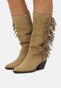 Botas altas de gamuza color beige con detalles de flecos, puntera puntiaguda y tacón apilado. La textura es suave con líneas limpias y uniformes.