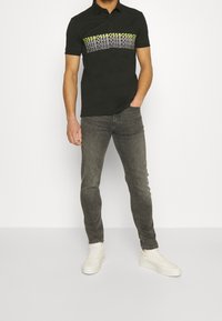 Svart polo skjorta med en grafisk band som har upprepande text i vitt och neon gult, matchad med grå jeans och vita sneakers.
