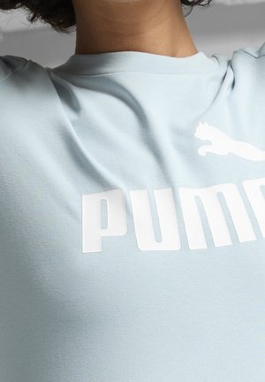 Persona con una camisa azul claro con el logo blanco de Puma y texto en el pecho, mostrando el torso superior y el cuello.