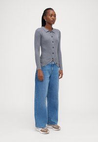 Sivoba ribana kardigana s ovratnikom in gumbom, v kombinaciji s širokimi modrimi denim hlačami. Obleka ima ozek kroj na vrhu.