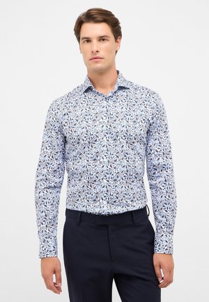 Chemise à manches longues blanche avec un motif floral bleu. Le tissu présente des motifs complexes avec des contours foncés et un col structuré.