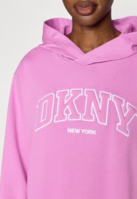 Roosa kapuutsiga pusa, valmistatud pehmest kangast, millel on valge tikandiga "DKNY" logo ja "NEW YORK" tekst. Sisaldab ruumikat kapuutsi ja pikki varrukaid.