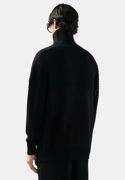 Uomo con capelli corti e scuri che indossa occhiali neri e un maglione a collo alto nero a coste, visto di spalle su uno sfondo uniforme.