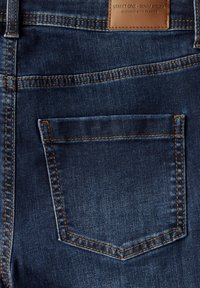 Jeans en denim bleu foncé avec une poche arrière unique, des coutures contrastantes brunes et une étiquette en cuir près de la taille.
