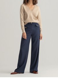 Beige V-halstrik en hooggetailleerde marineblauwe broek met brede pijpen, versierd met gouden knoopaccenten en zijzakken, met een gladde stoftextuur.