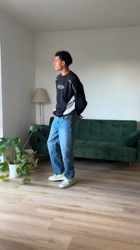 Svart og hvit sportslig genser, lyseblå jeans og hvite sneakers. Står nær en grønn sofa og en potteplante på tregulv.