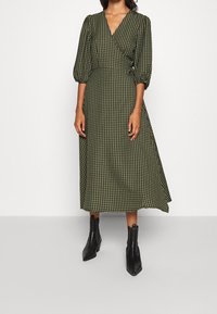 Robe portefeuille à carreaux verts avec des manches bouffantes, longueur mi-mollet et corsage ajusté. Associée à des bottines noires.