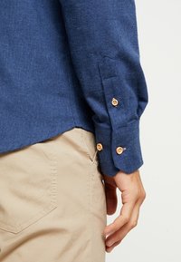 Mörkblå långärmad skjorta med knäppte manschetter, tillverkad av strukturerad väv. Ljusbeige byxor med en diskret fickdetalj.