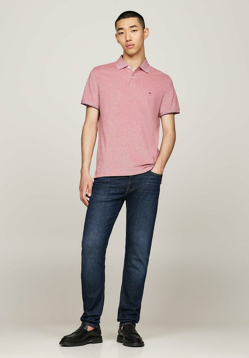 Rotes Poloshirt mit kontrastierendem Kragen, kombiniert mit dunkelblauen Jeans und schwarzen Loafers. Das Model steht mit einer Hand in der Tasche.