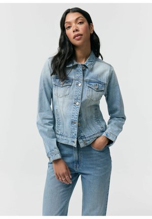 Kobieta z długimi ciemnymi włosami, ubrana w jasnoniebieską kurtkę jeansową i dopasowane dżinsy, stojąca z jedną ręką w kieszeni na jednolitym tle.