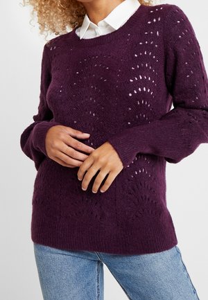 Vrouw die een paarse gebreide trui met een kanten patroon draagt over een wit overhemd met kraag en een blauwe spijkerbroek, handen zachtjes samengevouwen ter hoogte van de taille.