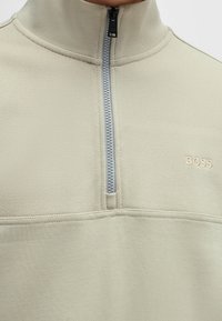 Ljust beige tröja med hög krage, med en silvrig dragkedja och präglad "BOSS"-logotyp. Texturerat tyg med kontrasterande paneldetalj.