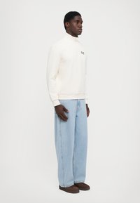 Hvit hettejakke med glidelås og høy krage, kombinert med lys blå vide jeans og brune sko. Glatt stofftekstur.