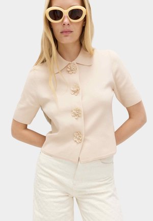 Kobieta w beżowej, krótkiego rękawa bluzce zapinanej na guziki w kształcie kwiatów, białych, teksturowanych spodniach oraz dużych, beżowych okularach przeciwsłonecznych.