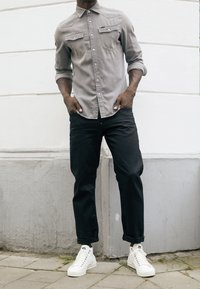 Camicia grigia con bottoni, due tasche sul petto e maniche arrotolate, abbinata a pantaloni neri con risvolti e sneakers alte bianche.