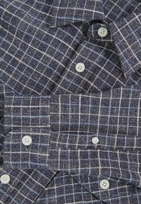 Cămașă cu carouri în nuanțe de bleumarin și albastru deschis, confecționată dintr-un material texturat. Are nasturi albi și un gât deschis. Modelele sunt distanțate uniform.