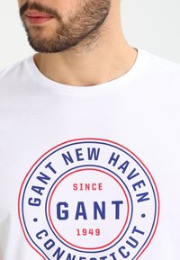 Camiseta blanca de algodón con un gráfico circular en rojo y azul que muestra el texto "GANT NEW HAVEN CONNECTICUT 1949" centrado en el pecho.