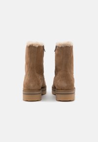 Un paio di stivali alla caviglia in suede marrone con bordo in shearling beige e suole in gomma, visti da dietro su uno sfondo bianco.