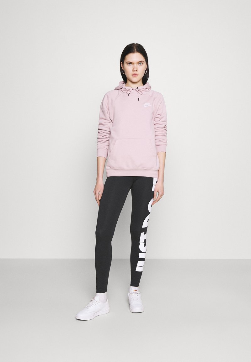 Helle rosa Kapuzenjacke mit langen Ärmeln und Fronttasche, kombiniert mit schwarzen Leggings, die mit weißem "JUST DO IT" Text bedruckt sind, sowie weißen Turnschuhen.