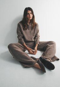 Kobieta z długimi brązowymi włosami siedząca na podłodze przy białej ścianie, ubrana w luźny brązowy sweter z dzianiny i spodnie oraz czarne buty.