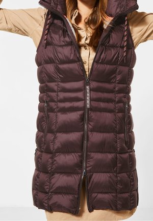 Femme portant une veste matelassée sans manches de couleur violet foncé avec fermeture éclair avant, par-dessus une chemise beige à manches longues.