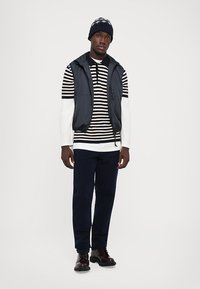 Mann in einem gestreiften schwarz-weißen Langarmshirt, schwarzem Weste, blauen Hosen, dunklen Lederschuhen und einer blauen Mütze, der vor einem weißen Hintergrund steht.