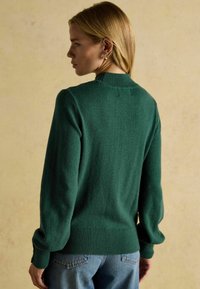 Tume roheline villane sviiter ribitud kaela- ja äärekangaga; kergelt puhvitatud pikkade varrukatega; kantud koos siniste teksadega. Esitletud tagantvaates.