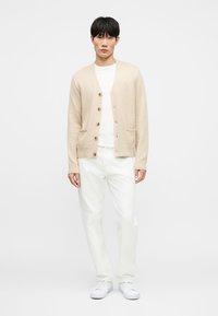 Νεαρός άνδρας που φοράει μπεζ ζακέτα πάνω από λευκό μπλουζάκι, λευκό παντελόνι και λευκά αθλητικά παπούτσια, στέκεται μπροστά σε απλό λευκό φόντο.