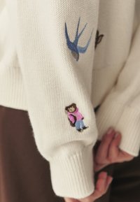 Sweter z kremowym splotem z haftowanymi detalami: niebieski jaskółka i niedźwiedź w różowo-niebieskich ubraniach na rękawach; ściągacze na mankietach.