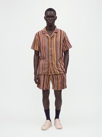 Jeune homme portant une chemise à manches courtes boutonnée et un short rayé assorti avec des baskets roses et des chaussettes noires sur fond blanc.