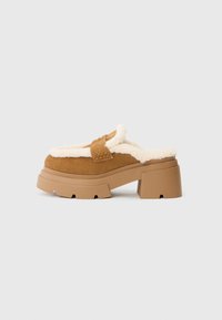 Scarpe slip-on in camoscio marrone con una morbida fodera color crema, suola piatta spessa e tacco a blocco. Presenta una fascia cucita sulla parte superiore.