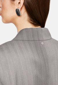 Gray herringbone-patterned fabric with a structured collar. Accompanied by a large black earring with a curved design and silver accents.