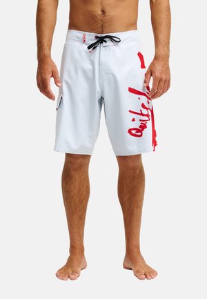 Mężczyzna w białych kąpielówkach do kolan z czerwonym logo Quiksilver na prawej nodze i czarnym sznurkiem.