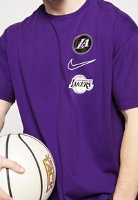 Camiseta de algodón morada con los logotipos de Los Angeles Lakers y Nike. Mangas cortas, cuello redondo, sostenida junto a un balón de baloncesto con superficie texturizada.