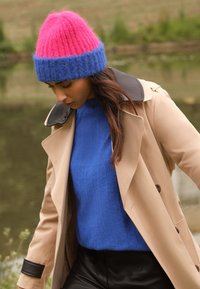 Multifarvet strikket hue med lyserød top og blå kant; hyggelig blå sweater; beige trenchcoat med sort krave; sorte figursyede bukser.