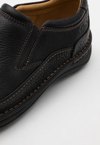Schwarzer Leder-Schlüpfschuh mit strukturiertem Finish, kontrastierenden braunen Nähten und runder Zehenform. Weiche Innenpolsterung sichtbar.