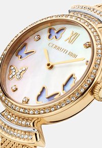 Reloj en tono dorado con esfera de nácar, presenta motivos de mariposas y acentos de cristal. Elegante caja redonda con una correa texturizada.