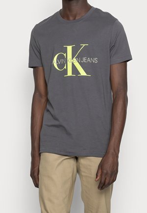 Mørkegrå Calvin Klein Jeans t-shirt med stort gult "cK" logo båret af person med mørk hud og beige bukser, stående mod en hvid baggrund.