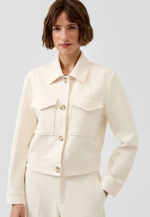 Vrouw met kort bruin haar, gekleed in een crèmekleurig jasje met knopen en klepzakken, en bijpassende broek, staand met één hand in de zak.