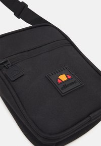 Black fabric crossbody bag featuring a zip closure, logo patch with red and yellow accents, and an adjustable strap. Textured surface.