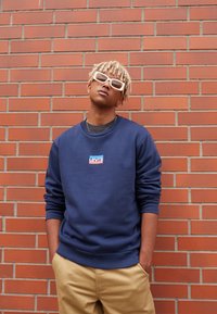 Mörkblå sweatshirt med rundad hals, med en Levi's logotyp påsydd. Bärs med beige byxor; accessoarer med ljusa solglasögon.