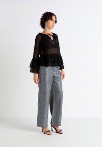 Black sheer long-sleeve blouse with ruffled hem, paired with gray striped wide-leg trousers. Model wears black open-toe heels with embellishments.