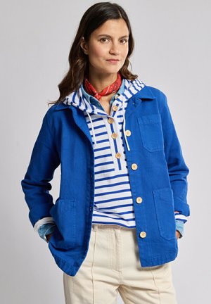 Femme portant une veste bleue sur un sweat à capuche rayé bleu et blanc, un pantalon beige et un foulard rouge autour du cou, debout les mains dans les poches.