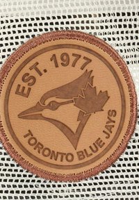 Leather patch featuring a blue jay and maple leaf, embossed text “EST. 1977 TORONTO BLUE JAYS,” on a textured white mesh background.