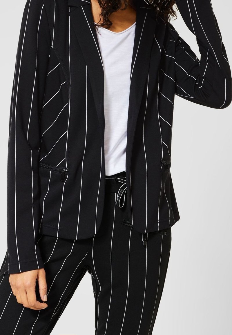 Blazer noir à rayures blanches verticales, design à un bouton, deux poches zippées, tissu doux, complété par un T-shirt blanc en dessous.