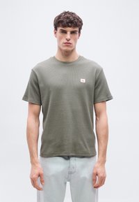 Νέος άνδρας με κοντά σγουρά μαλλιά, φοράει μια ελιάς πράσινη υφασμάτινη μπλούζα με μικρό κόκκινο λογότυπο και ανοιχτό μπλε τζιν, στέκεται μπροστά σε απλό φόντο.