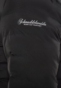Juodas pūkuotas švarkas su siuvinėjimu. Ant jo yra balta siuvinėta užrašas "Schmuddelwedda WENN DAT PLATTERT." Minkšta, glotni tekstūra.